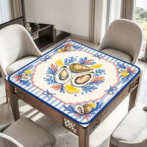 May include: Square table mat with a nautical theme. The mat features a cream-colored center with watercolor illustrations of oysters, lemon slices, and blue coral. A blue and white striped border with rope accents frames the design.