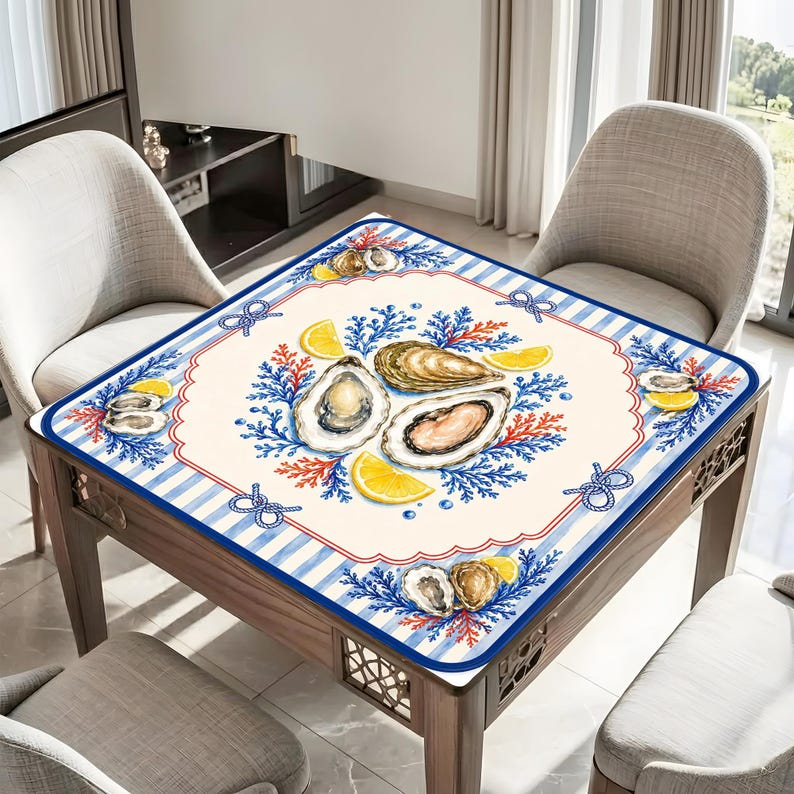 May include: Square table mat with a nautical theme. The mat features a cream-colored center with watercolor illustrations of oysters, lemon slices, and blue coral. A blue and white striped border with rope accents frames the design.