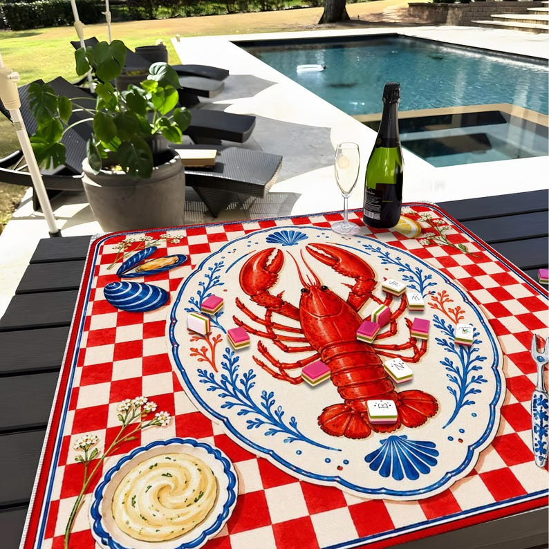 Op de afbeelding: Een rood-wit geruite placemat met een kreeftenillustratie op een decoratieve schaal. De schaal is omgeven door blauwe bloemmotieven. Een fles champagne en een glas staan op de tafel.