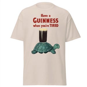May include: A beige t-shirt with a graphic of a turtle carrying a glass of Guinness beer. The text on the shirt reads "Have a GUINNESS when you're TIRED."