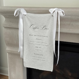 May include: A linen coffee bar sign with white ribbon ties. The sign lists latte flavors and add-ons, including vanilla, caramel, hazelnut, pistachio, lavender, and cane sugar. It also features an affogato option with espresso and vanilla bean ice cream.