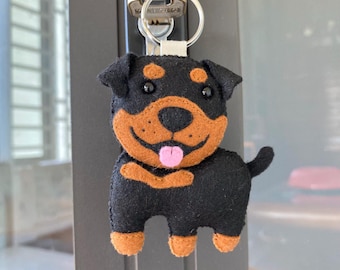 Op de afbeelding: Een vilten sleutelhanger van een Rottweiler-hond. De sleutelhanger heeft een zwarte body met bruine markeringen op het gezicht, de borst en de poten. De hond heeft een lachende uitdrukking met een roze tong. Een zilveren sleutelring en een witte stoffen lus zijn bevestigd.