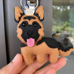 Puede incluir: Un llavero de fieltro hecho a mano con forma de perro pastor alemán. El perro tiene un cuerpo marrón con marcas negras, ojos negros y una lengua rosa. El llavero está sujeto a un anillo de metal plateado.