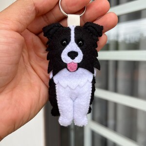 May include: A felt keychain shaped like a Border Collie dog with black and white fur, a pink tongue, and black bead eyes. The dog figure is attached to a silver metal split ring by a small white fabric loop, featuring a layered design.