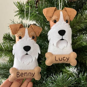 Puede incluir: Dos adornos de fieltro con forma de cabeza de terrier con barbas blancas y orejas de color canela. Cada adorno tiene una base en forma de hueso con los nombres Benny y Lucy bordados en hilo marrón, colgados de cordones blancos.