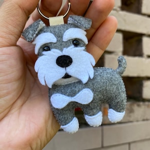 May include: A grey felt Schnauzer dog keyring with white detailing on the face, paws, and a bow tie. The keyring has a gold-coloured ring and a beige fabric loop. The dog has a black nose and eyes.