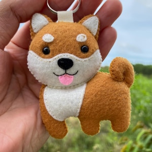 Puede incluir: Llavero de fieltro hecho a mano con forma de perro Shiba Inu. El llavero presenta un perro marrón y blanco con una lengua rosa, ojos negros y un llavero de metal. El perro está sonriendo y tiene una cola esponjosa.