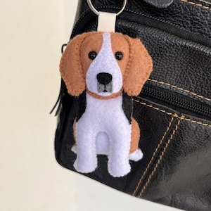 Puede incluir: Un llavero de fieltro con forma de perro beagle en colores marrón, blanco y negro. El perro tiene orejas caídas, una mancha blanca en la cara y una nariz negra. Está sujeto a una anilla de metal plateado mediante una pequeña trabilla de tela blanca.