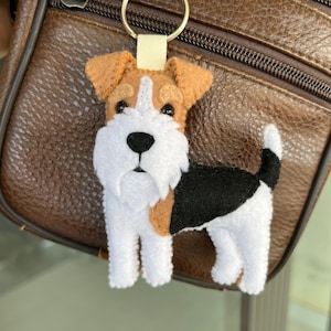 May include: A felt Wire Fox Terrier dog keyring. The dog is white, black, and brown, with a white muzzle and chest, brown ears, and a black tail. The keyring is attached to a metal ring and a beige fabric loop.