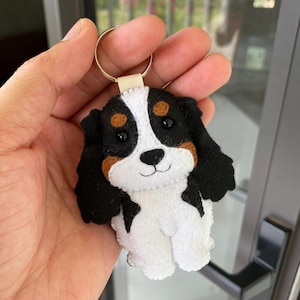 Op de afbeelding: Een handgemaakte sleutelhanger van een Cavalier King Charles Spaniel. De pluche hond heeft een witte vacht, zwart-bruine aftekeningen en een gouden sleutelring. Het gezicht van de hond heeft zwarte ogen en een gestikte glimlach.