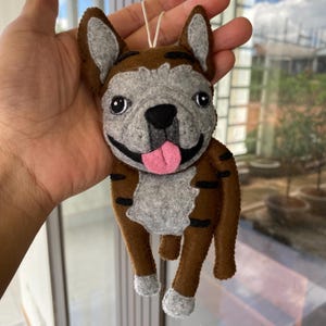 May include: A handmade felt ornament in the shape of a French Bulldog. The dog has a brown body with black stripes, gray face and ears, and a pink tongue. The ornament is designed with black eyes and a smiling expression.