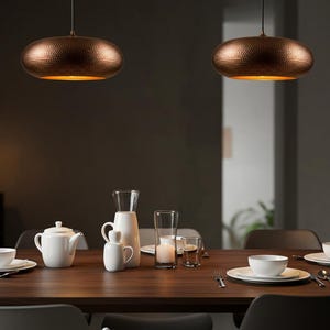 May include: Two copper-colored pendant lights hang above a wooden dining table set with white dishes, a teapot, and glassware. The lights have a textured surface and a warm glow, illuminating the dining area. The scene suggests a modern, elegant dining room.