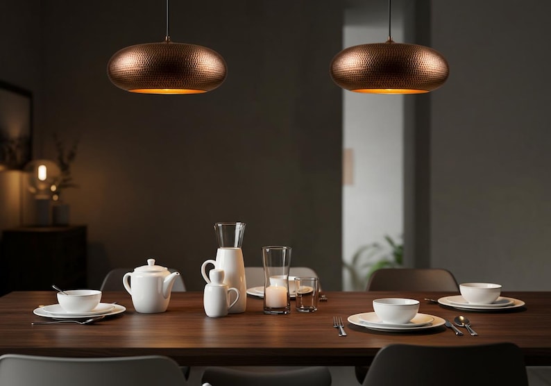 May include: Two copper-colored pendant lights hang above a wooden dining table set with white dishes, a teapot, and glassware. The lights have a textured surface and a warm glow, illuminating the dining area. The scene suggests a modern, elegant dining room.