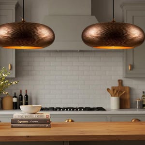 May include: Two copper-colored pendant lights with a hammered texture hang above a wooden kitchen island. The lights have a warm, golden interior. The kitchen features gray cabinets, a white tile backsplash, and a vase of greenery.