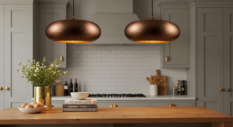 May include: Two copper-colored pendant lights with a hammered texture hang above a wooden kitchen island. The lights have a warm, golden interior. The kitchen features gray cabinets, a white tile backsplash, and a vase of greenery.