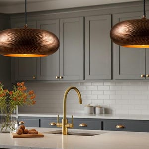 May include: Two copper-colored pendant lights with a hammered texture hang above a kitchen island. The lights have a warm, golden glow from the interior. The kitchen features gray cabinets, a white countertop, and a gold faucet.