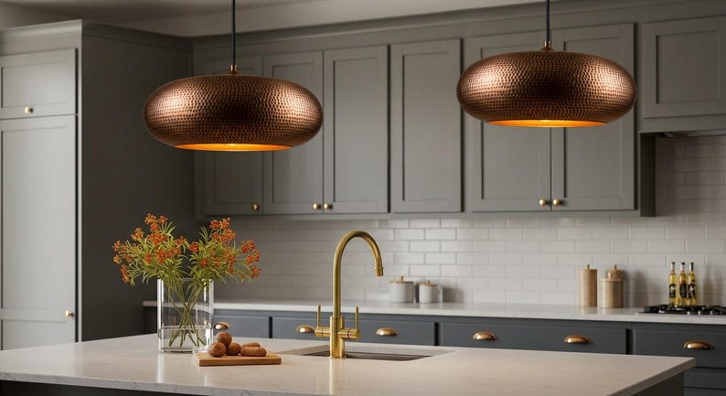 May include: Two copper-colored pendant lights with a hammered texture hang above a kitchen island. The lights have a warm, golden glow from the interior. The kitchen features gray cabinets, a white countertop, and a gold faucet.