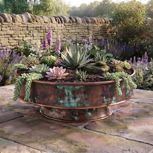 May include: A round, low-profile copper planter with a weathered, verdigris patina finish. The planter features a pedestal base and is filled with a variety of succulents, including a central agave, a pink echeveria, and trailing sedum plants.