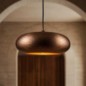 May include: A copper-colored, hammered metal pendant light fixture. The light has a rounded, oval shape and hangs from a black cord. The interior of the light fixture glows with a warm, golden light. The background features an arched doorway.