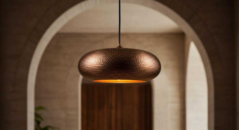 May include: A copper-colored, hammered metal pendant light fixture. The light has a rounded, oval shape and hangs from a black cord. The interior of the light fixture glows with a warm, golden light. The background features an arched doorway.