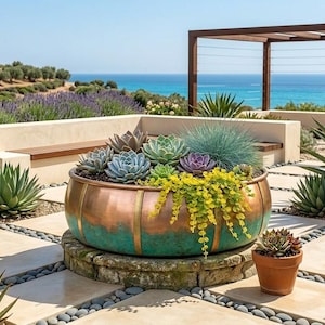 Op de afbeelding: Een grote koper- en turkooiskleurige plantenbak, gevuld met vetplanten en hangplanten, staat op een stenen voet. De scène omvat agave, lavendel, uitzicht op de zee en een pergola op de achtergrond.