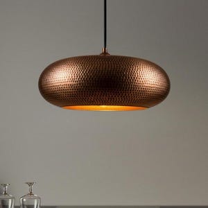 May include: A copper-colored, oval-shaped pendant light fixture with a textured, hammered surface. The interior of the light fixture has a warm, golden glow. The light hangs from a black cord, and is a decorative lighting element.