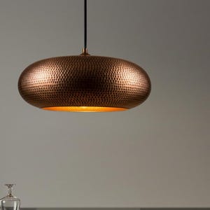 May include: A copper-colored, oval-shaped pendant light with a textured surface. The light fixture hangs from a black cord and emits a warm, golden glow from its interior. This decorative lighting fixture is a stylish addition to any home.
