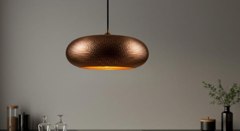 May include: A copper-colored, oval-shaped pendant light with a textured surface. The light fixture hangs from a black cord and emits a warm, golden glow from its interior. This decorative lighting fixture is a stylish addition to any home.