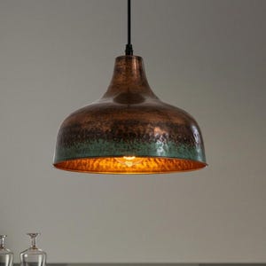 Puede incluir: Una lámpara colgante de color cobre con una superficie texturizada y un borde verde-azul. La lámpara cuelga de un cable negro e ilumina con un brillo cálido. La lámpara es una adición elegante a cualquier hogar.