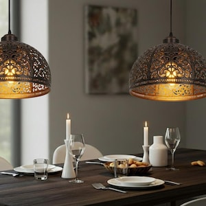 K&ouml;nnte beinhalten: Zwei verzierte Pendelleuchten in Bronze h&auml;ngen &uuml;ber einem Esstisch. Die Lampen haben ein detailliertes, durchbrochenes Design und werfen warmes Licht. Der Tisch ist mit Kerzen, Tellern und Gl&auml;sern gedeckt, was eine einladende Atmosph&auml;re schafft.