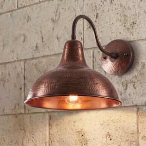 May include: A wall-mounted light fixture featuring a wide, dome-shaped shade with a hammered copper finish. The lamp includes a curved arm connecting the shade to a circular wall plate, with a warm light bulb visible inside the metallic dome.