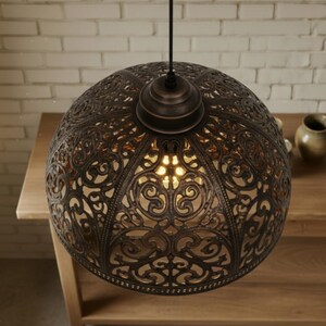 K&ouml;nnte beinhalten: Verzierte, bronzefarbene Pendelleuchte mit kugelf&ouml;rmigem Design. Die Leuchte hat ein kompliziertes, durchbrochenes Muster, das Licht durchscheinen l&auml;sst. Die Leuchte h&auml;ngt an einem schwarzen Kabel und beleuchtet einen Holztisch.
