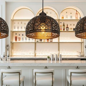 K&ouml;nnte beinhalten: Drei bronzefarbene Pendelleuchten mit aufwendigen, durchbrochenen Mustern h&auml;ngen &uuml;ber einer Bar. Die Leuchten strahlen ein warmes, goldenes Licht aus. Die Bar hat wei&szlig;e Arbeitsplatten, wei&szlig;e St&uuml;hle und eine Ausstellung von Flaschen im Hintergrund.