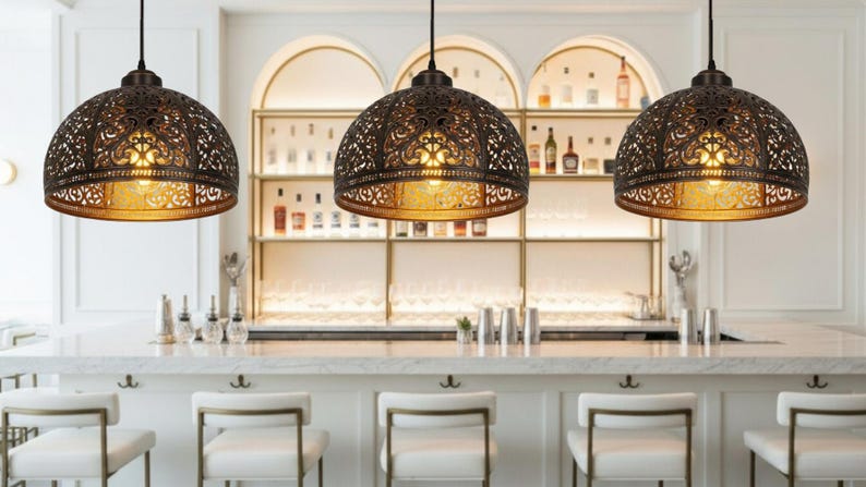 K&ouml;nnte beinhalten: Drei bronzefarbene Pendelleuchten mit aufwendigen, durchbrochenen Mustern h&auml;ngen &uuml;ber einer Bar. Die Leuchten strahlen ein warmes, goldenes Licht aus. Die Bar hat wei&szlig;e Arbeitsplatten, wei&szlig;e St&uuml;hle und eine Ausstellung von Flaschen im Hintergrund.