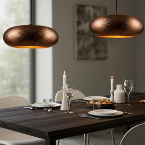 May include: Two copper-colored pendant lights with a hammered texture hang above a dark wood dining table. The lights have a warm, golden interior. The table is set with white plates, glasses, and candles, creating a cozy dining atmosphere.