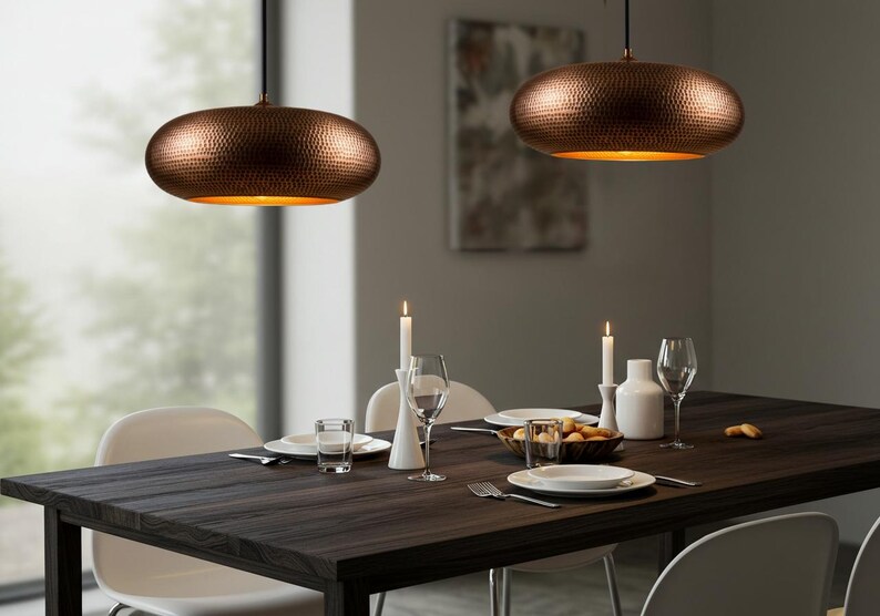 May include: Two copper-colored pendant lights with a hammered texture hang above a dark wood dining table. The lights have a warm, golden interior. The table is set with white plates, glasses, and candles, creating a cozy dining atmosphere.