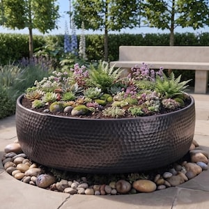 Puede incluir: Una jardinera grande, redonda y de metal marrón oscuro con textura martillada, llena de una variedad de pequeñas suculentas verdes y moradas y musgo. La jardinera se asienta sobre un lecho circular de piedras de río lisas y multicolores.
