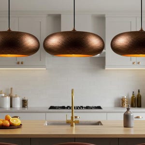 May include: Three copper-colored pendant lights with a hammered texture hang above a kitchen island. The lights have a warm, golden glow emanating from within. The kitchen features white cabinets, a wooden island, and a gold faucet.