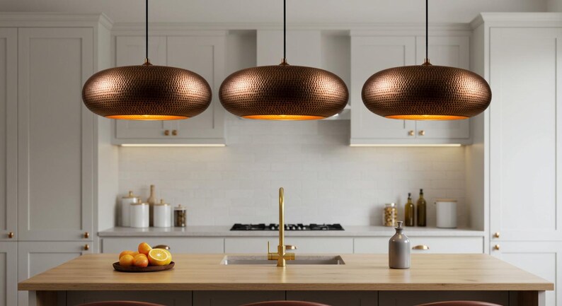 May include: Three copper-colored pendant lights with a hammered texture hang above a kitchen island. The lights have a warm, inviting glow. The kitchen features white cabinets, a wooden island, and a gold faucet.