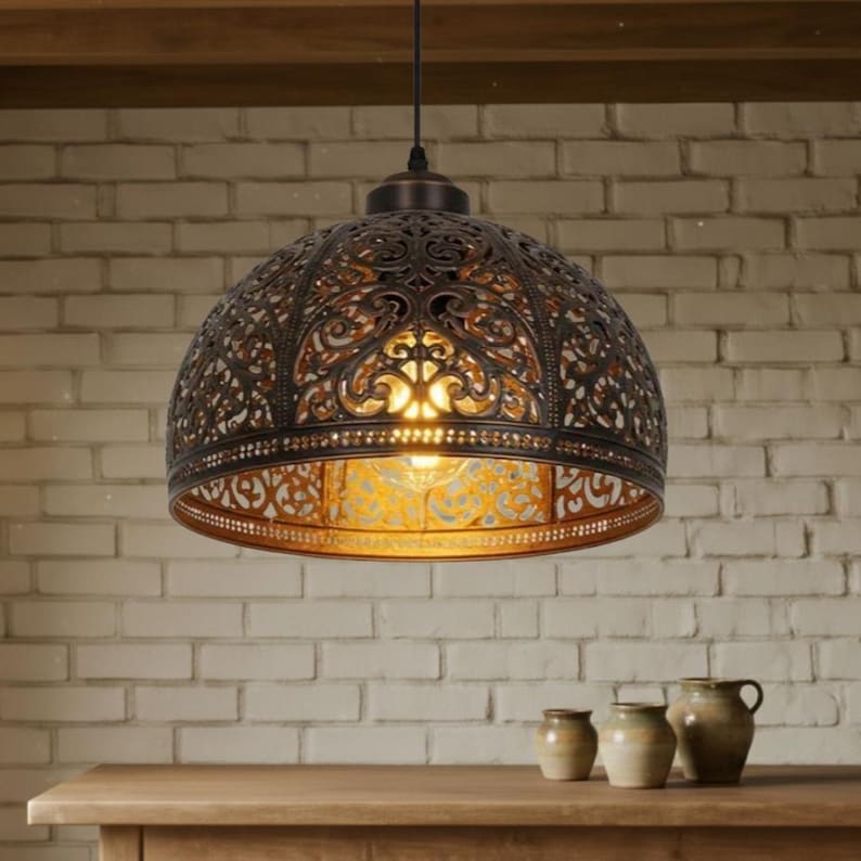 May include: Ornate, bronze-colored pendant light with a filigree design, hanging from a dark cord. The light fixture is dome-shaped, with a warm glow emanating from within. The background features a brick wall and wooden table.