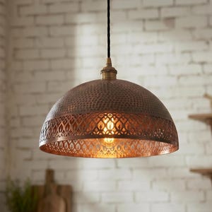 Pode incluir: Uma luminária pendente de cor cobre com uma cúpula martelada e um intrincado design vazado na parte inferior. A luminária pende de um cabo preto e é iluminada por dentro, projetando um brilho quente. Está suspensa em frente a uma parede de tijolos brancos.
