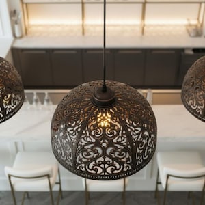 K&ouml;nnte beinhalten: Drei bronzefarbene Pendelleuchten mit aufwendigen, durchbrochenen Mustern h&auml;ngen &uuml;ber einer wei&szlig;en Arbeitsplatte und Barhockern. Die Leuchten strahlen ein warmes Licht aus, das die detaillierten Muster hervorhebt. Die K&uuml;che hat dunkle Schr&auml;nke und eine wei&szlig;e Marmorarbeitsplatte.