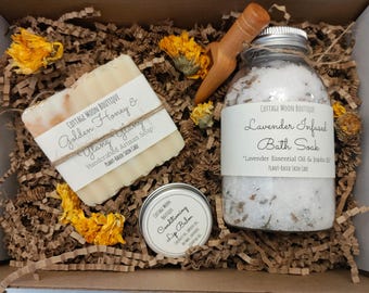 May include: A gift box containing a bar of Golden Honey & Ylang Ylang soap, a jar of Lavender Infused Bath Soak, and a small tin of Conditioning Lip Balm. The box includes a wooden scoop and dried yellow flowers.