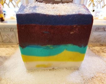 May include: A rectangular bar of handmade soap with layered colors: white, yellow, teal, burgundy, and navy blue. The top is sprinkled with white crystals. The soap is sitting on a surface covered in white crystals.