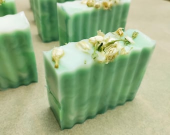 May include: A close-up of a green and white bar of soap with white flower petals on top. The soap has a rippled texture.