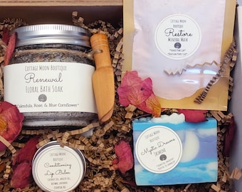 May include: A wooden box filled with self-care products. It includes a jar of floral bath soak, a wooden scoop, a mineral mask, a lip balm tin, and a bar of soap. The products are surrounded by dried flowers and paper filler.