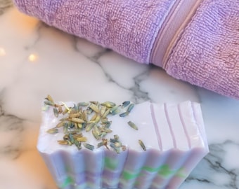 May include: A lavender-colored bar of soap with dried lavender buds on top. The soap has a scalloped edge and vertical lines on the top. A folded lavender towel rests in the background. The soap is on a marble surface.
