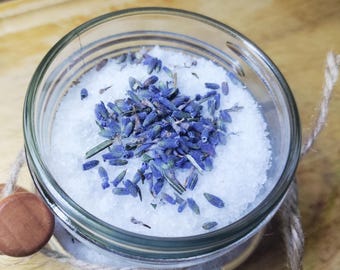May include: A clear glass jar filled with white bath salts and topped with dried lavender buds. The jar is tied with twine and has a wooden accent. The jar sits on a wooden surface.