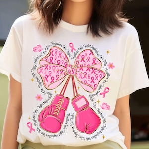 May include: White t-shirt featuring a pink bow with pink ribbons and boxing gloves, and the text "Together We Fight". The design includes pink ribbons, butterflies, and stars, symbolizing breast cancer awareness.