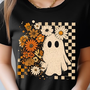 Puede incluir: Camiseta negra con un diseño gráfico de fantasma con un diseño floral y a cuadros. El fantasma es de color crema con ojos negros, rodeado de flores naranjas, amarillas y blancas. Un patrón de cuadros está en el lado derecho.